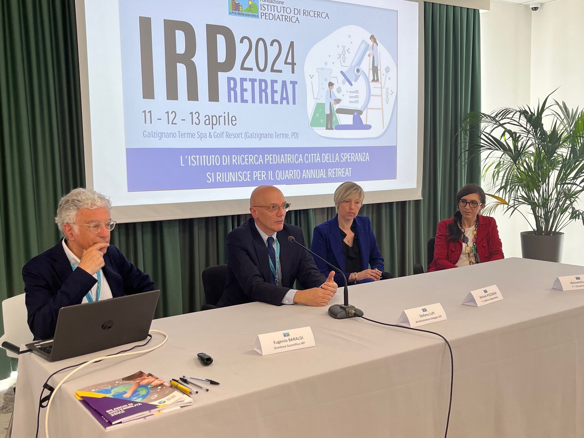 Una relatrice parla al microfono davanti a una platea, all’interno dell’Istituto di Ricerca Pediatrica "Città della Speranza". Sullo sfondo è visibile un grande schermo con una presentazione proiettata. L’evento si svolge in un ambiente formale, con partecipanti seduti e attenti.