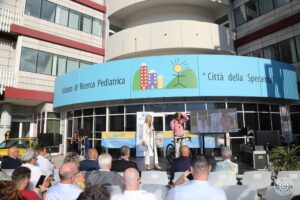 Sul palco due persone che parlano al microfono al pubblico sedute davanti a loro a un evento davanti all'ingresso di Città della Speranza